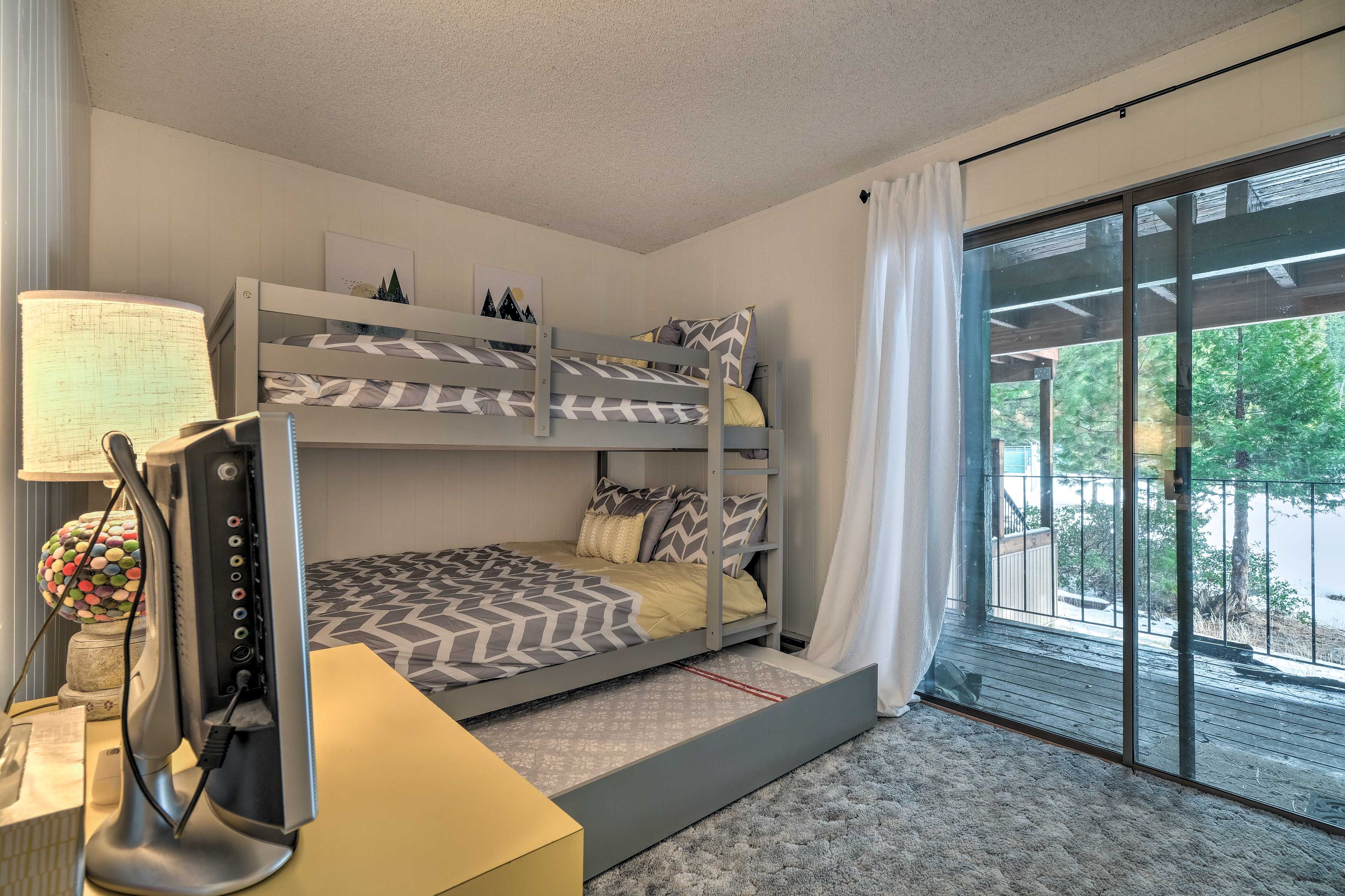 A bunk bed room with a desk and lamp, a large window with a view of trees, and a ceiling fan.