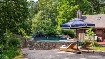 A stone pool with lounge chairs and an umbrella, surrounded by lush greenery and a wooden cabin.