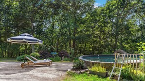 A pool area with a lounge chair and umbrella, surrounded by trees and a small pond. - Guilford Carriage House at Granite Lodge! (Guilford)