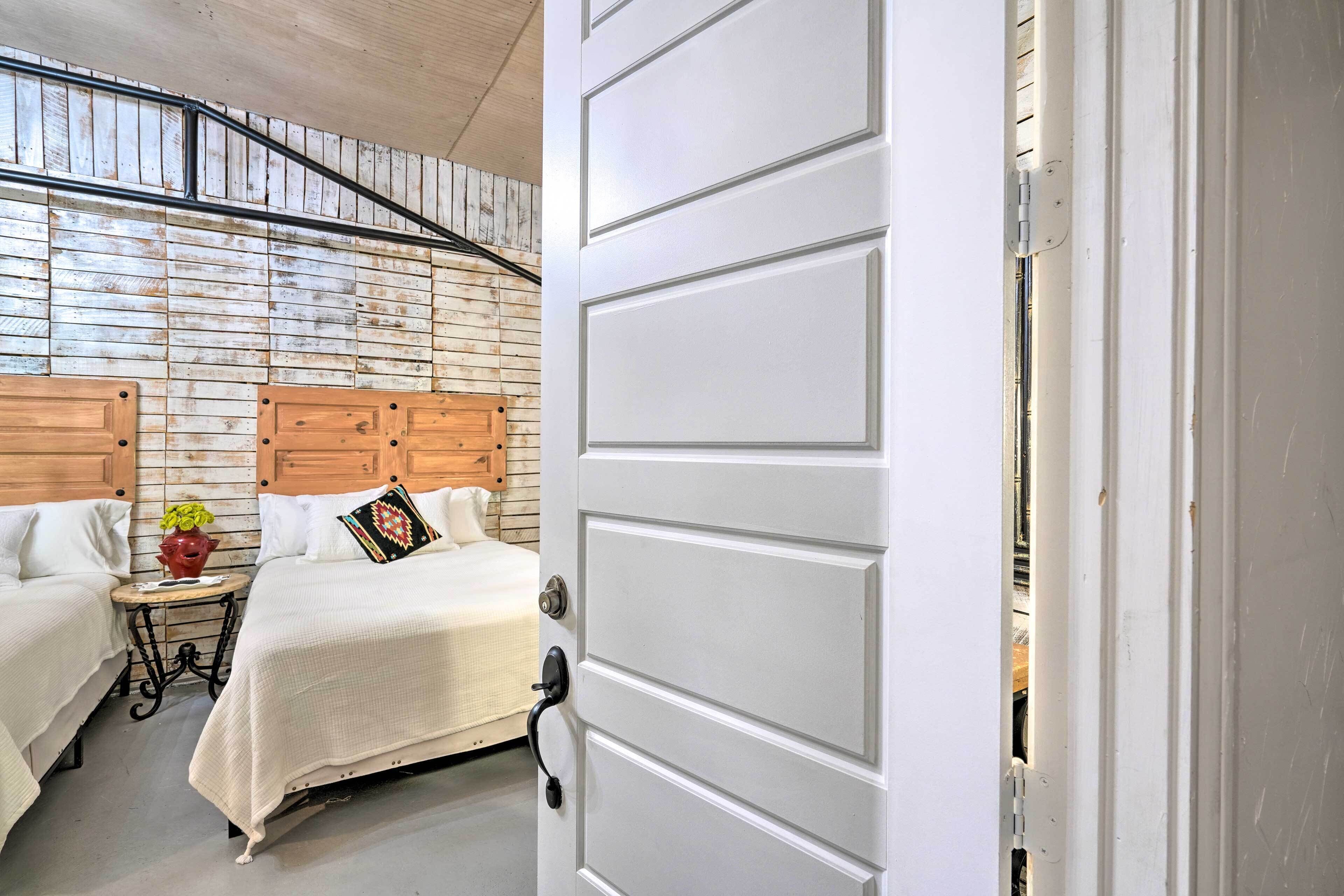 Un dormitorio con dos camas, un cabecero de madera, una mesita con un jarrón y una puerta blanca.