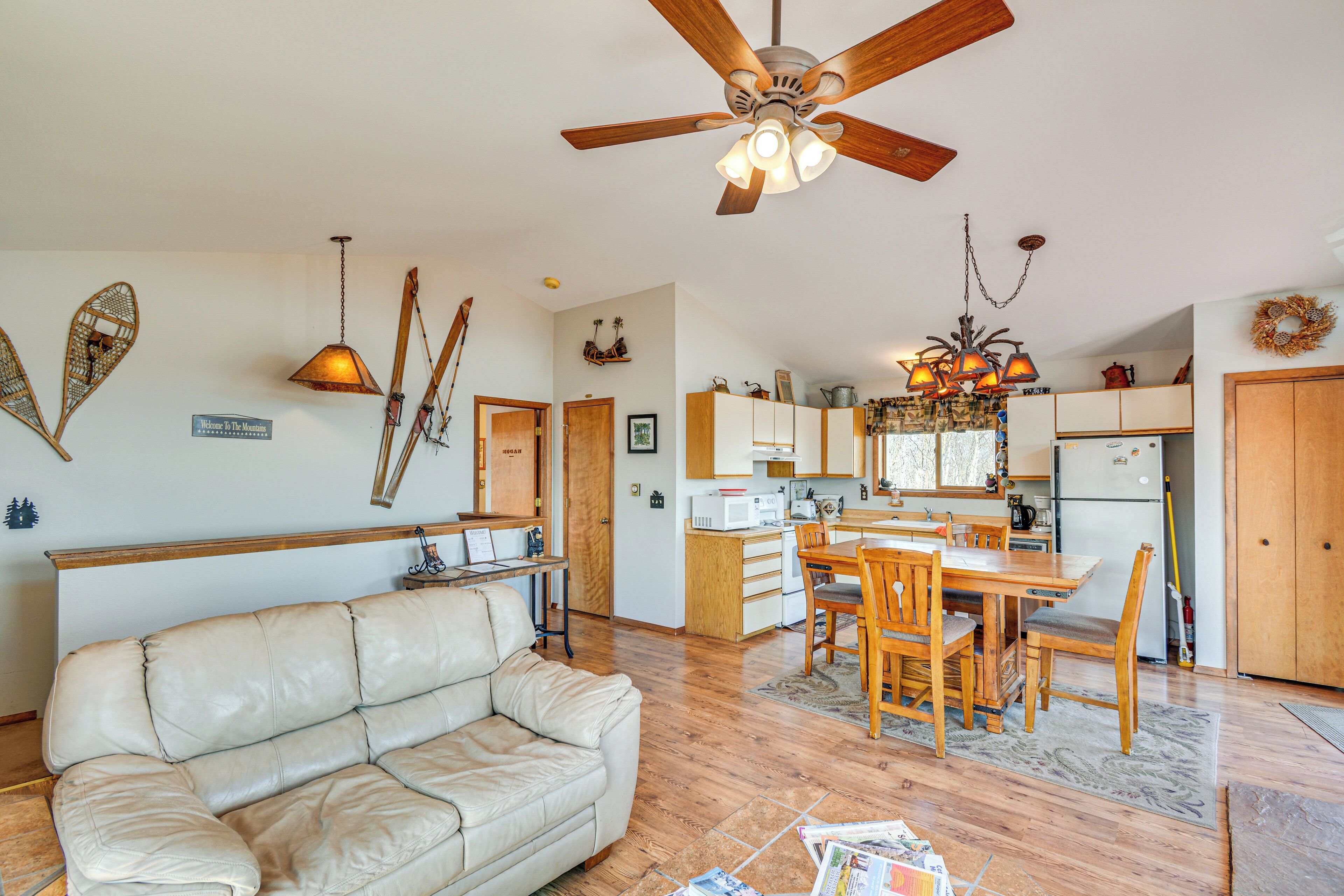 A spacious living area with a leather sofa, wooden dining table, and chairs, a kitchen with wooden cabinets and appliances, and a ceiling fan.