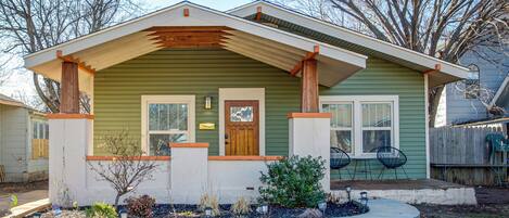 A single-story house with a green exterior, a front porch with wooden posts, a small front yard with plants and a fire pit, and a wooden fence in the background.