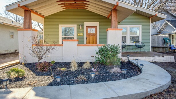 A single-story house with a green exterior and a wooden porch. There is a small garden with various plants and a pathway leading to the front door. - Chic Lawton Home, 5 Mins to Fort Sill! (Lawton)
