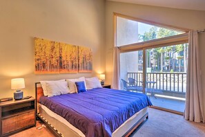A bedroom with a large bed, a nightstand with a lamp, a painting on the wall, and a balcony with a view of trees and buildings.