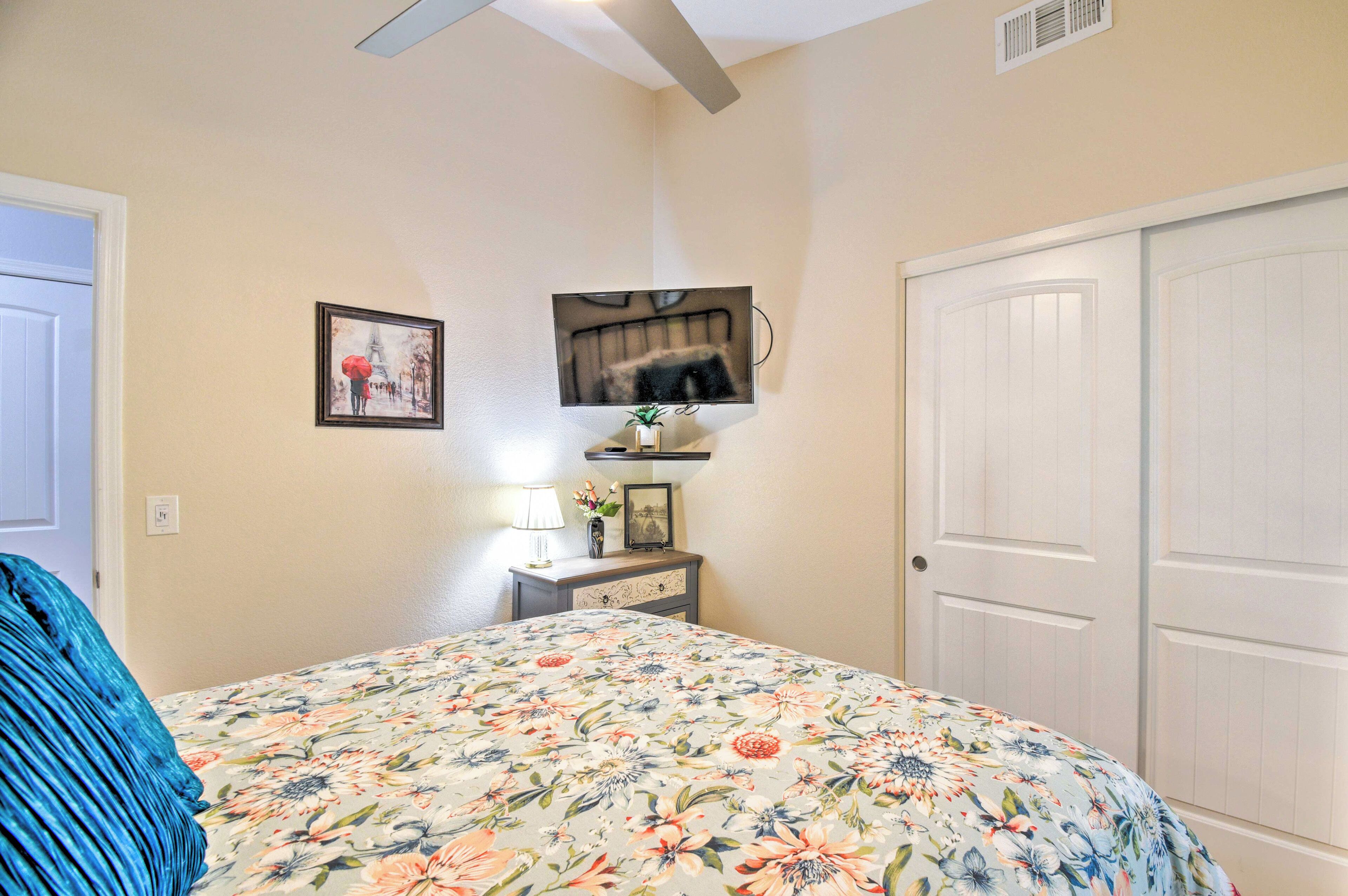 Une chambre avec une couvre-lit à motifs floraux, un téléviseur fixé au mur, une table de chevet avec une lampe et une porte sur le côté droit.