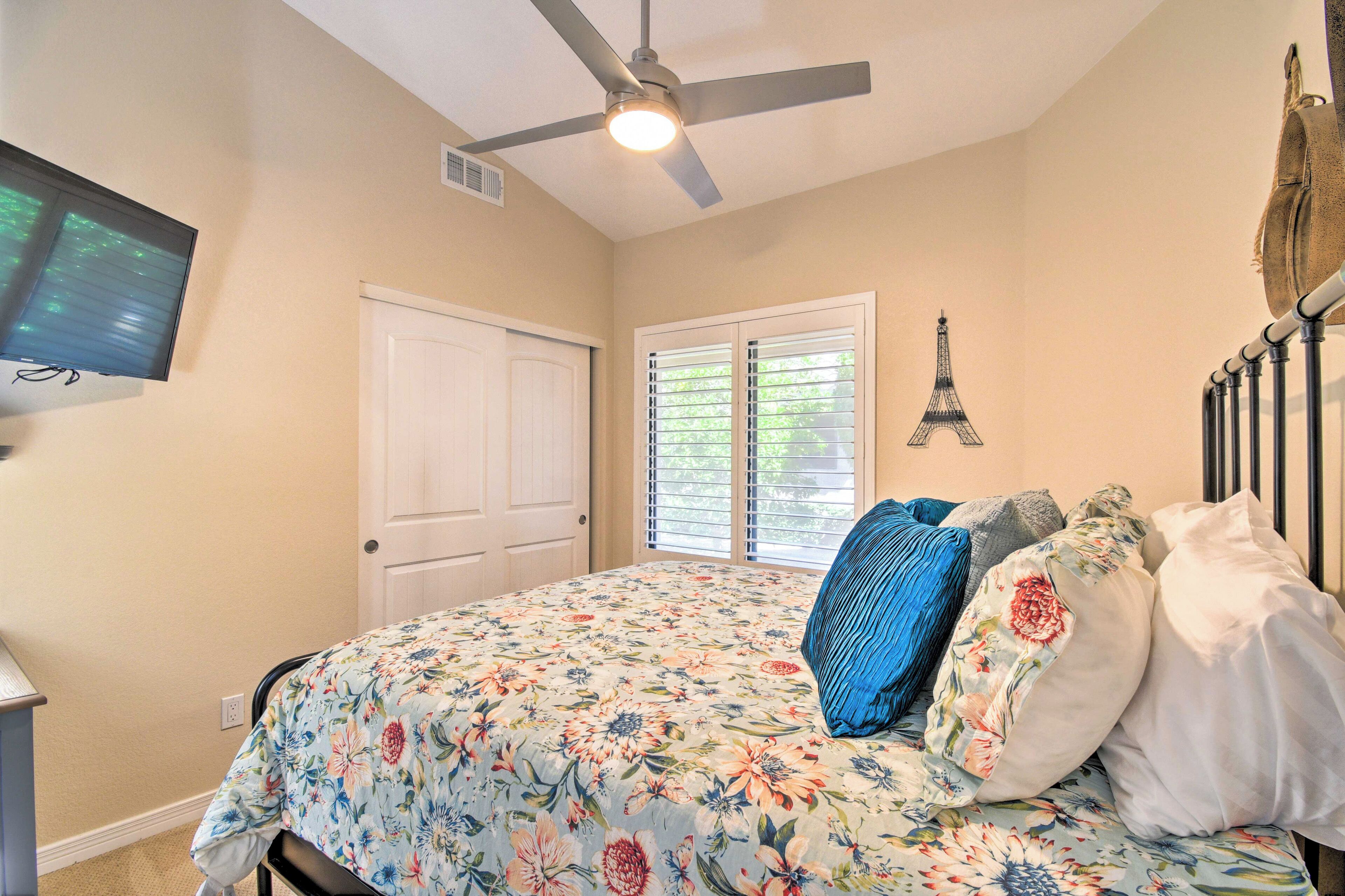 A bedroom with a bed, a ceiling fan, a TV, a door, and a window with blinds.