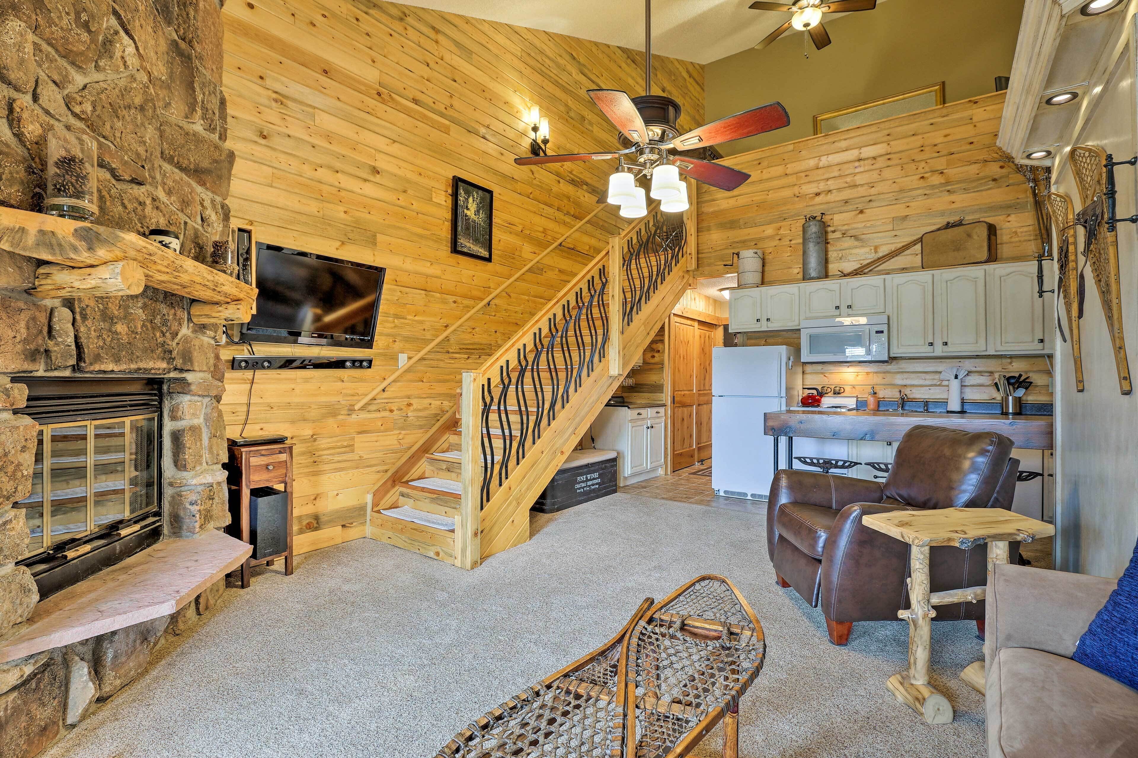 A log cabin interior with a stone fireplace, wooden staircase, kitchen area, and leather furniture.