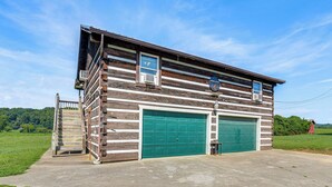 A two-story log cabin with a green garage door, a small attached shed, and a concrete driveway. - Rogersville Barn Apartment on 27 Acres w/ Pond! (Rogersville)