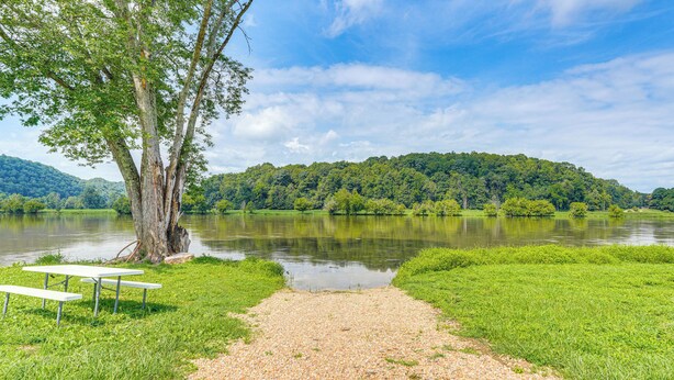A gravel path leading to a picnic area with benches, surrounded by lush greenery and a calm lake, with hills in the background. - Rogersville Barn Apartment on 27 Acres w/ Pond! (Rogersville)
