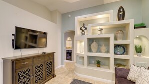 Un salon comprenant un téléviseur à écran plat, un meuble brun avec des portes en verre décoratives, une étagère blanche ornée d’objets décoratifs et un canapé brun agrémenté de coussins à motifs.