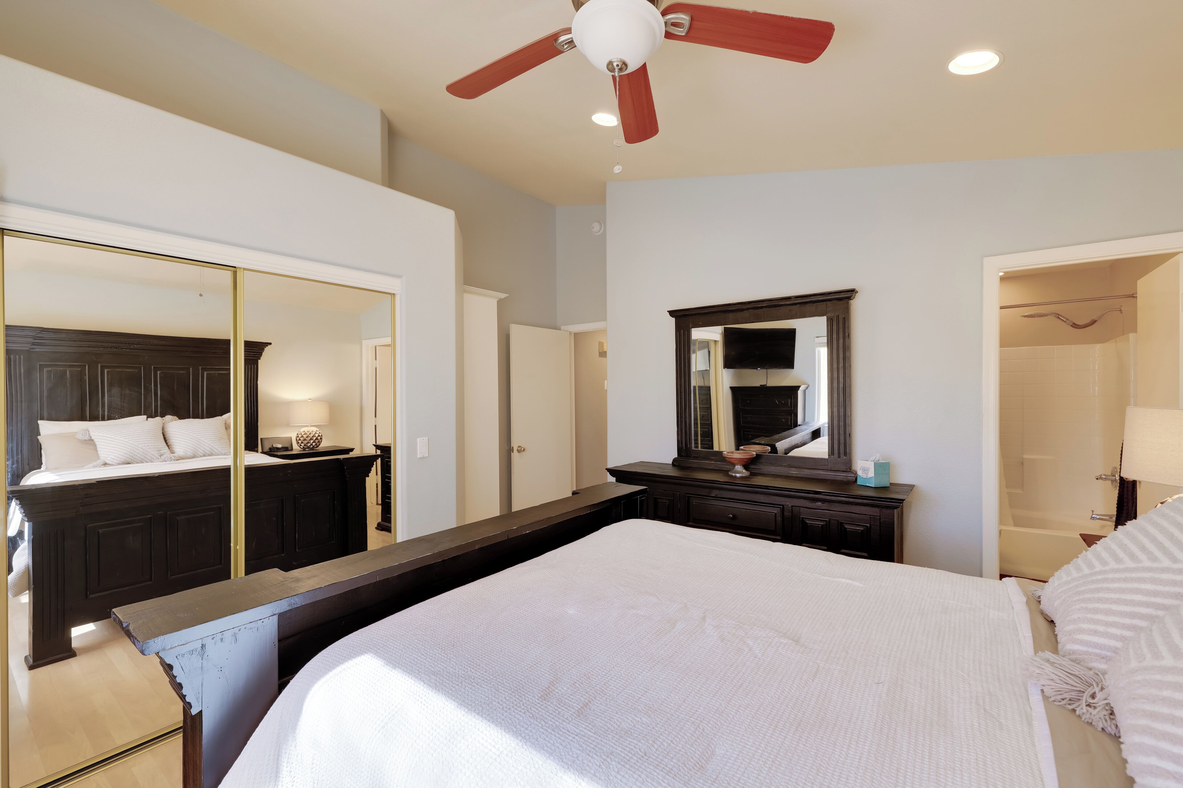 A bedroom with a large bed, a ceiling fan, a mirror, and a door leading to a bathroom.