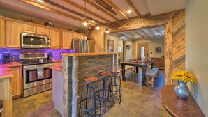 Una cocina rústica con vigas de madera, una isla central con taburetes, electrodomésticos de acero inoxidable y una zona de comedor con mesa y bancos de madera.