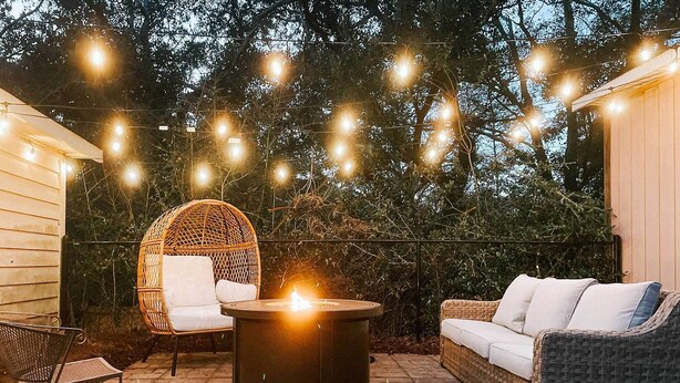 An outdoor patio with wicker furniture, a round table with a lit fire, and string lights.
