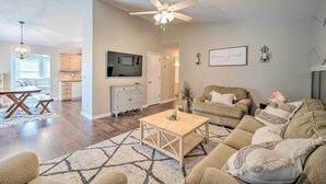 A spacious living room with a large sofa, a coffee table, and a ceiling fan. There is a kitchen area visible in the background.