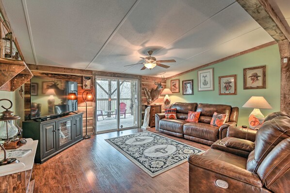 A spacious living room with a leather sofa, a television, and a large window overlooking a deck. - Rustic Country Escape - 20 Minutes From Nashville (Greenbrier)