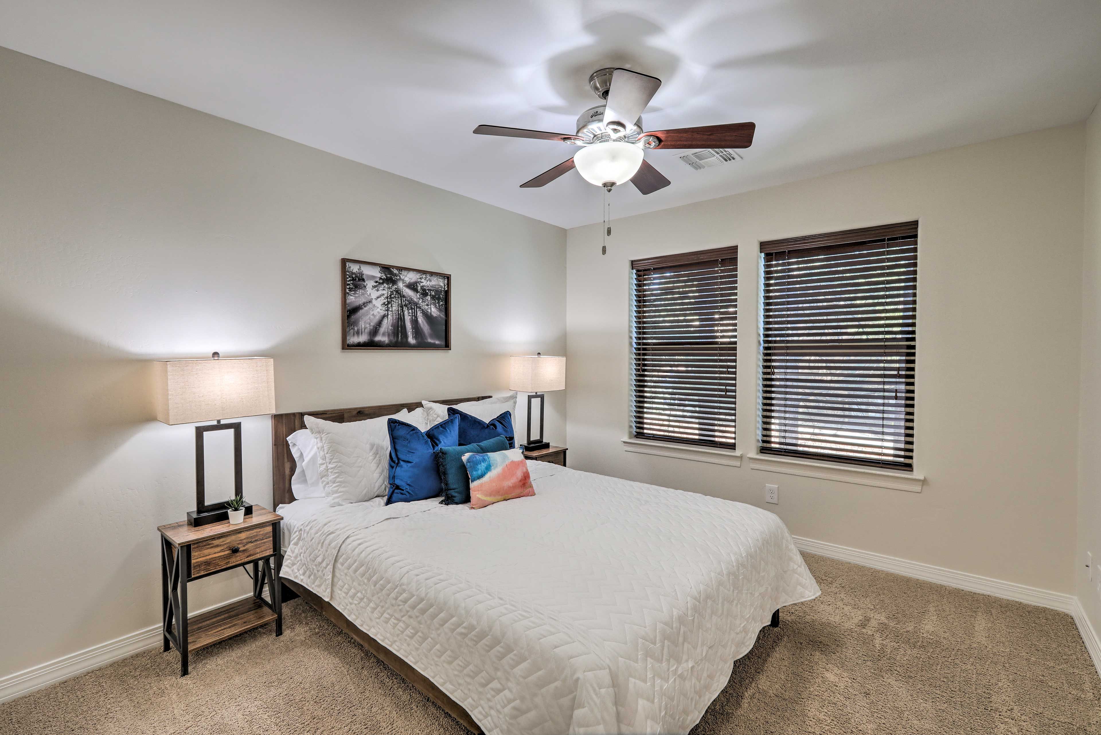 A bedroom with a bed, two bedside tables, a ceiling fan, a window with blinds, and a wall-mounted artwork.