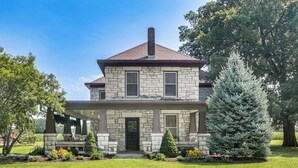 A two-story stone house with a covered porch, a chimney, and a well-maintained lawn with various shrubs and a large coniferous tree. - Pet-friendly Home: Grill & Fire Pit in Manhattan (Manhattan)