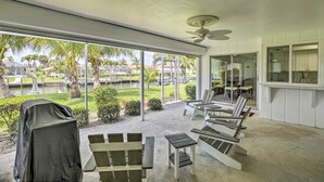 A covered patio with outdoor seating, a view of a landscaped area with palm trees, and a ceiling fan. - Bright Home w/ Dock & Canal View in Cape Coral! (Cape Coral)