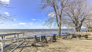 A lakeside picnic area with metal tables and chairs, a wooden deck, and a large tree.