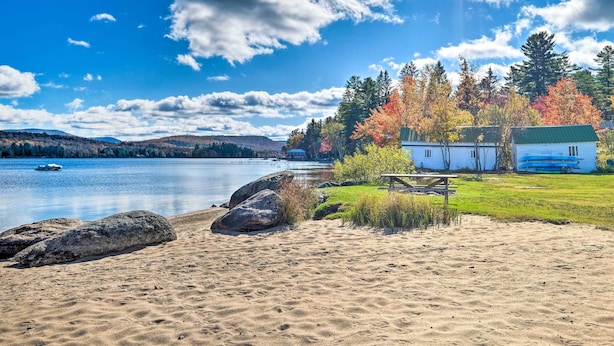 A lakeside scene with a sandy beach, a wooden bench, and a small cabin with a green roof. - Fire Pit & Beach: Lake Pleasant Home w/ Views (Speculator)