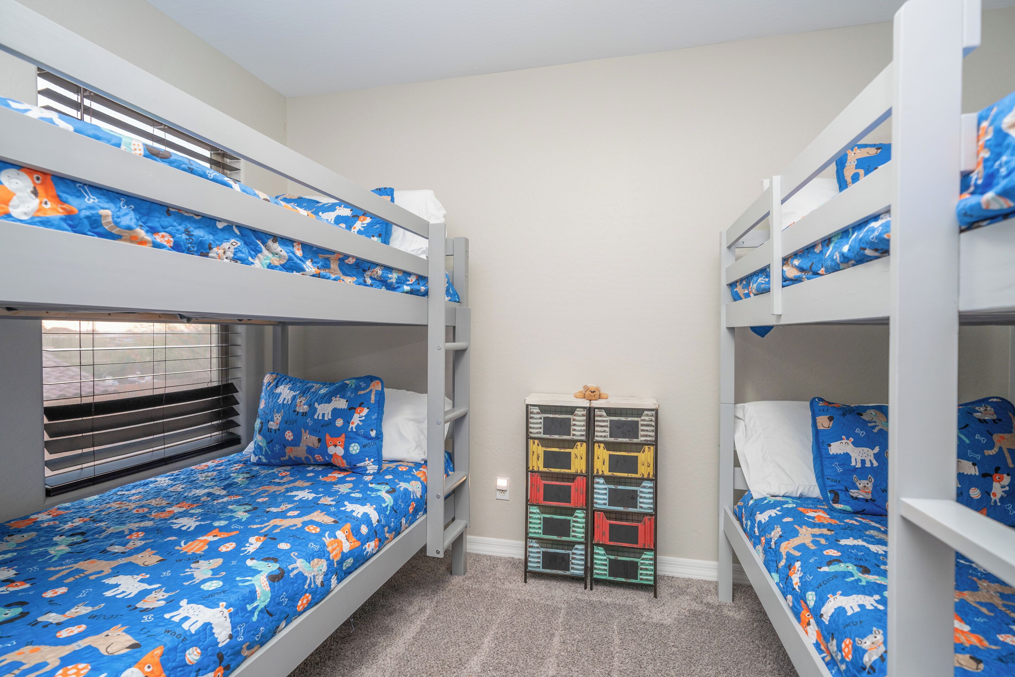 Habitación con literas, un edredón con motivos de animales en tonos azules, un mueble de almacenaje con compartimentos de colores y una ventana con persianas.