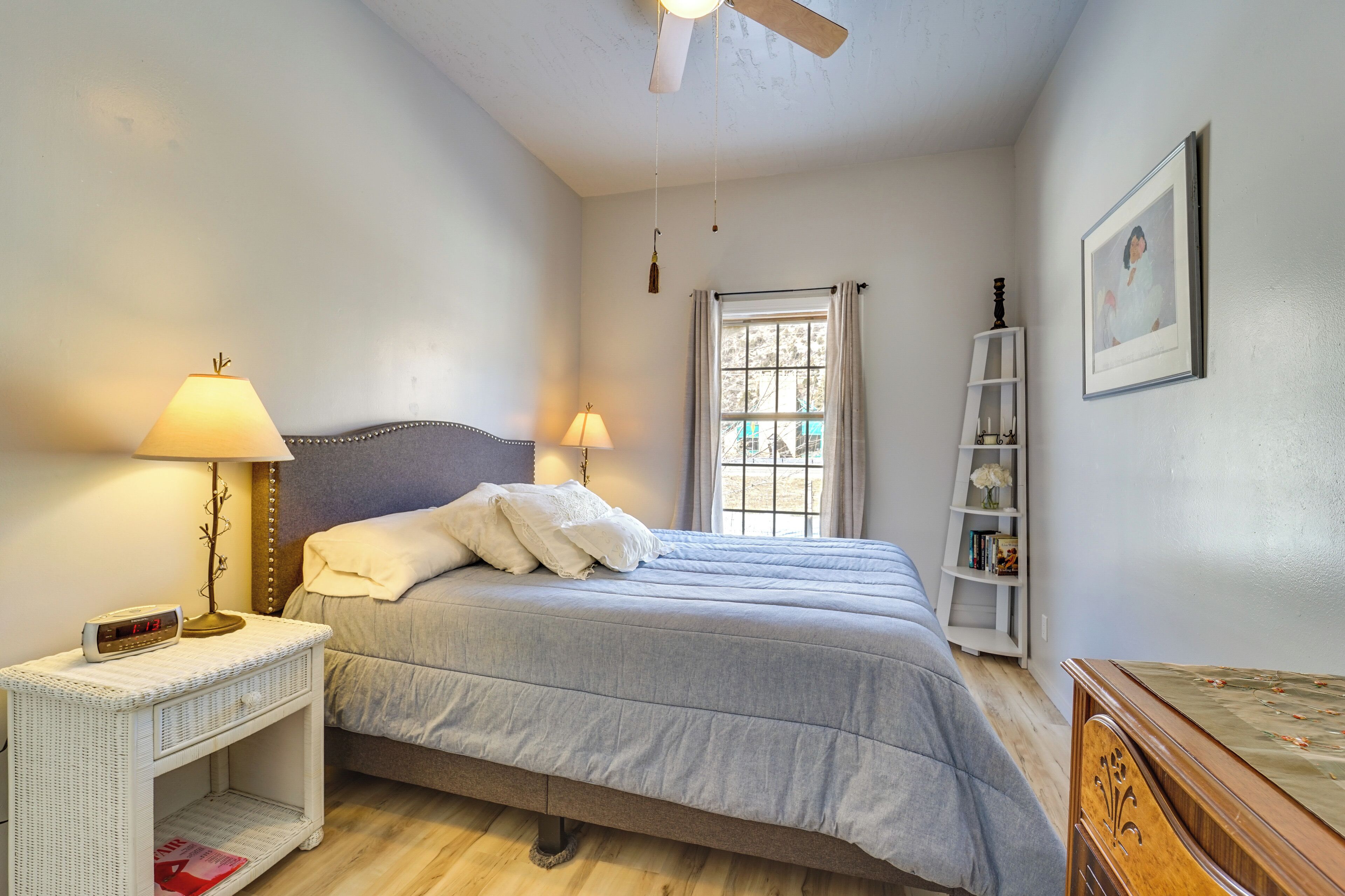 Un dormitorio con cama, lámparas de noche, mesita de noche, estantería, ventilador de techo y ventana con cortinas.
