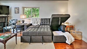 Ein gemütliches Wohnzimmer mit einem grauen Ecksofa, einem gemusterten Sessel, einem kleinen Beistelltisch mit einer Lampe und einem an der Wand befestigten Fernseher.