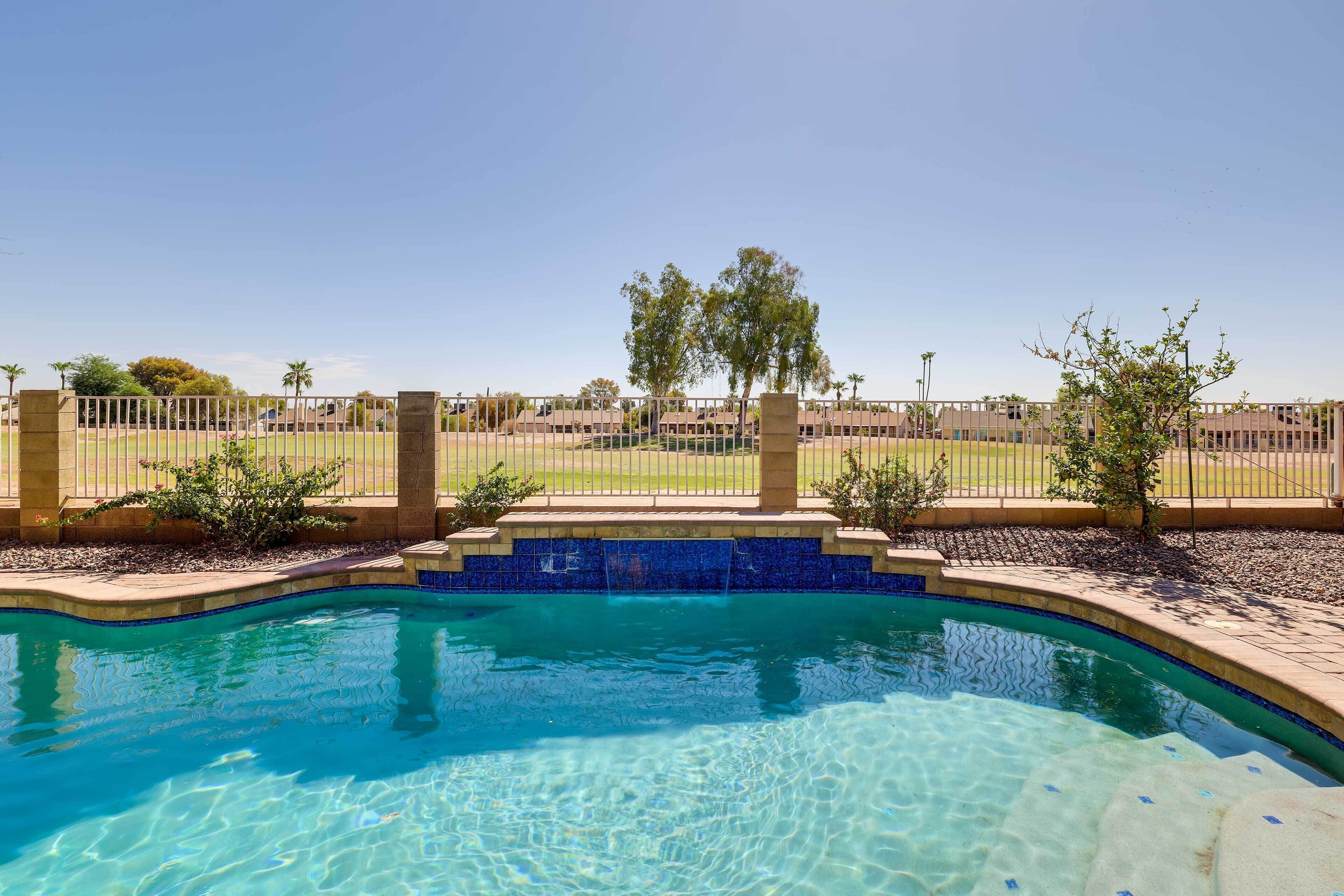 A clear swimming pool with a blue mosaic border, surrounded by a fence and a grassy area with trees.
