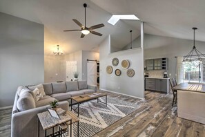 A spacious living room with a gray sofa, a wooden coffee table, and a patterned rug. There is a ceiling fan, a chandelier, and a dining area with a table and chairs.