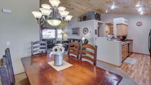 Een eetgedeelte met een houten tafel, stoelen en een kroonluchter. Achtergrond toont een keuken met kasten en een koelkast.