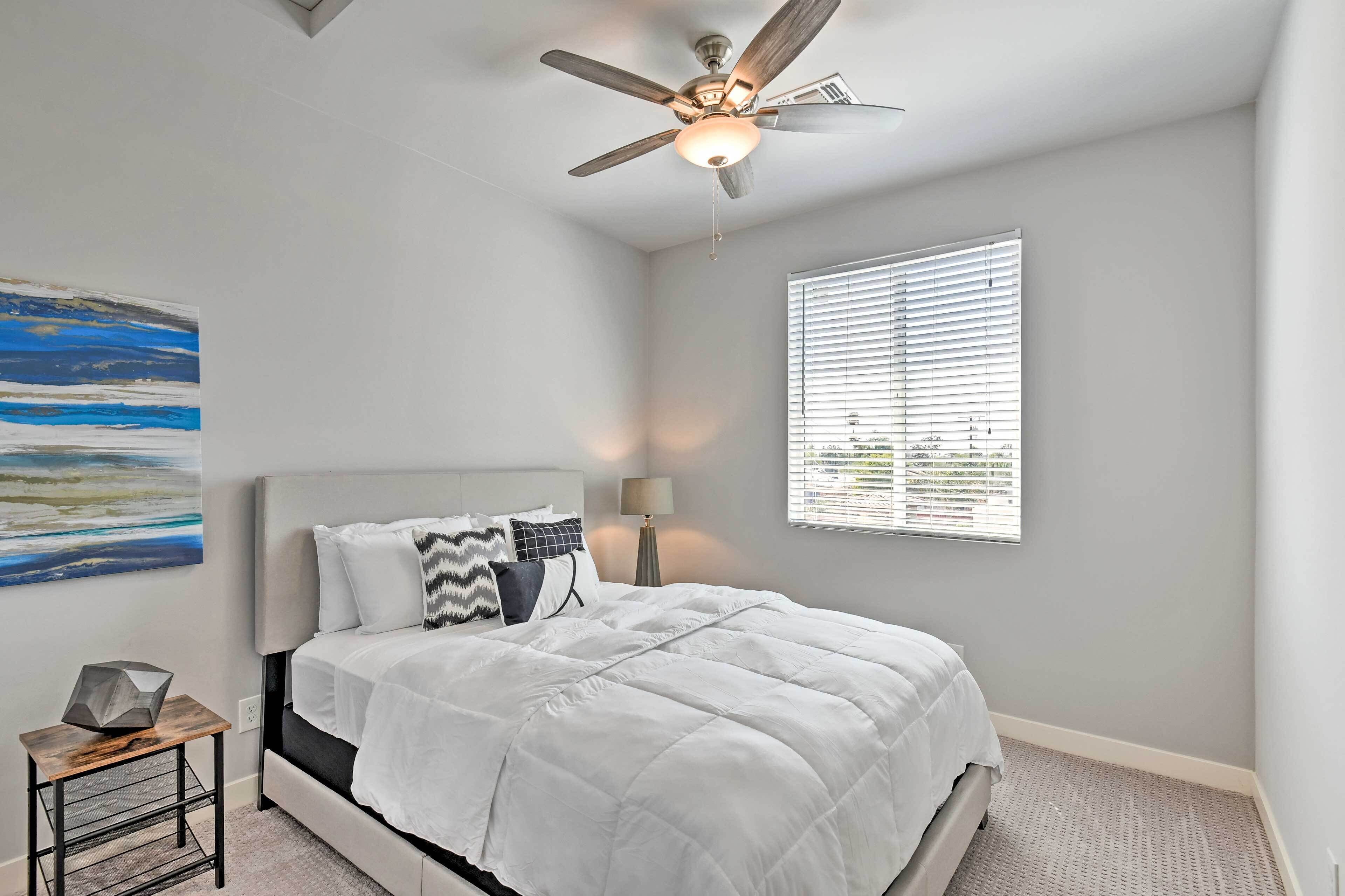 A bedroom with a bed, a ceiling fan, a window with blinds, a painting on the wall, and a small table with a lamp.