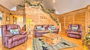 A cozy log cabin interior with a wooden staircase, leather armchairs, a wooden coffee table, and a Christmas tree.