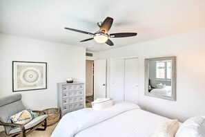 Ein Schlafzimmer mit einem Bett, einem Sessel, einer Kommode, einem Deckenventilator, einem gerahmten Spiegel und einem Wandbild.