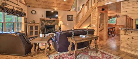 A spacious living room with a wooden ceiling, a fireplace, a large leather sofa, a wooden coffee table, and a red patterned rug.