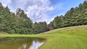 Paisagem tranquila com um lago, área gramada e uma cabana de madeira cercada por árvores densas.