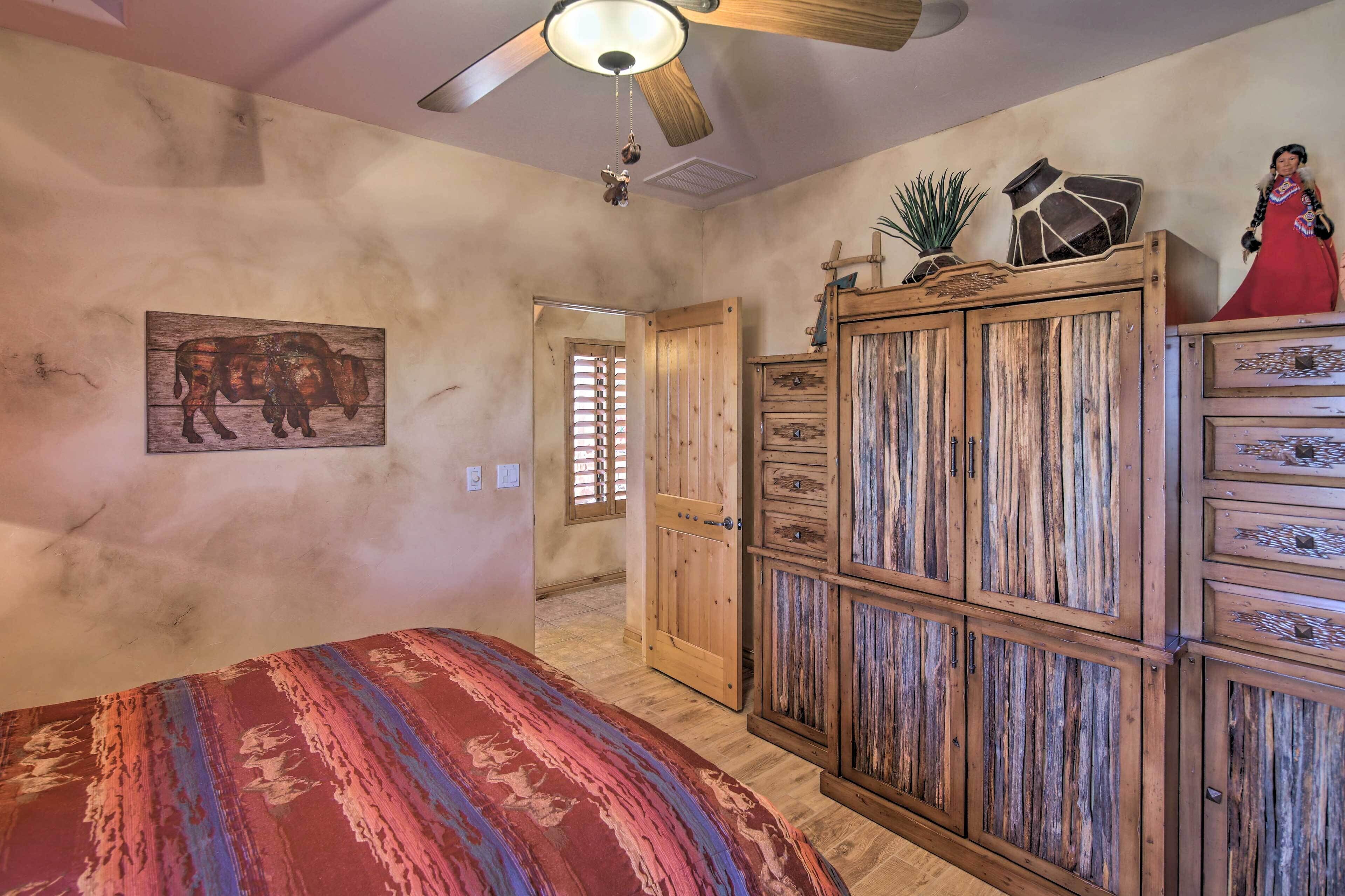 Um quarto com um guarda-roupa de madeira, uma cama com um cobertor colorido, um ventilador de teto e uma planta fixada na parede.