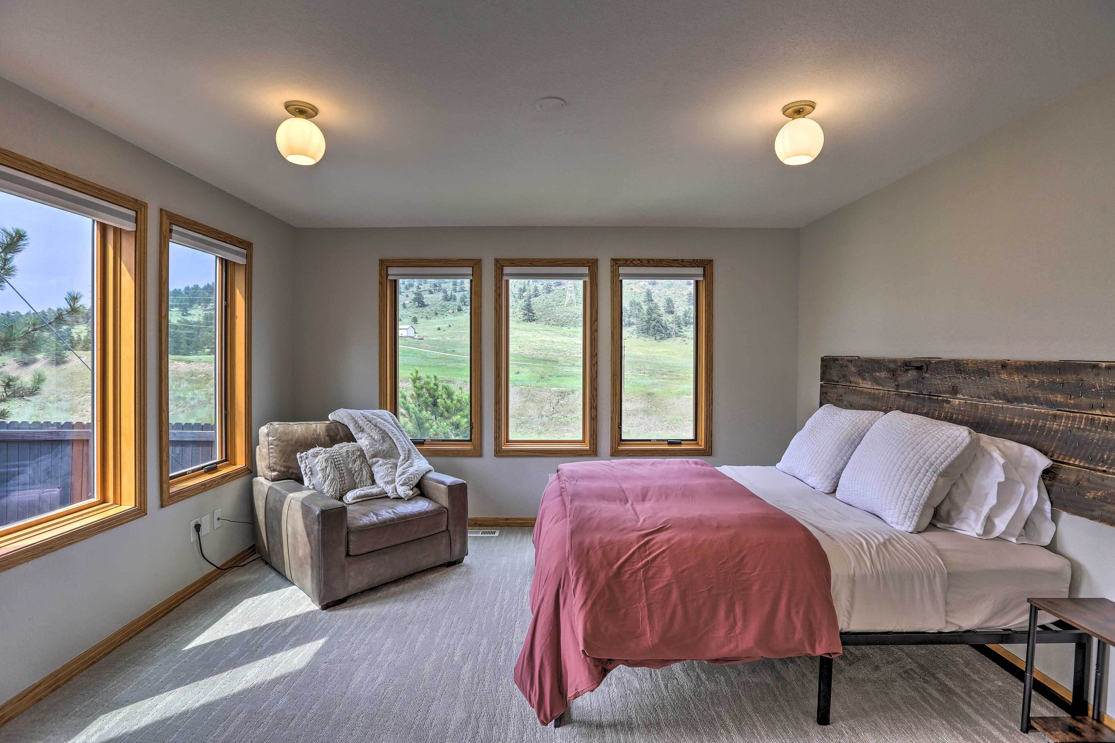 A bedroom with a bed, a chair, and large windows with a view of a green landscape.