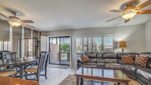 A spacious living room with a dark leather sofa, a wooden coffee table, and a dining table with chairs. There are ceiling fans and a large window with blinds. - Walk to Lake Havasu Beaches: Condo w/ Pool Access! (Lake Havasu City)