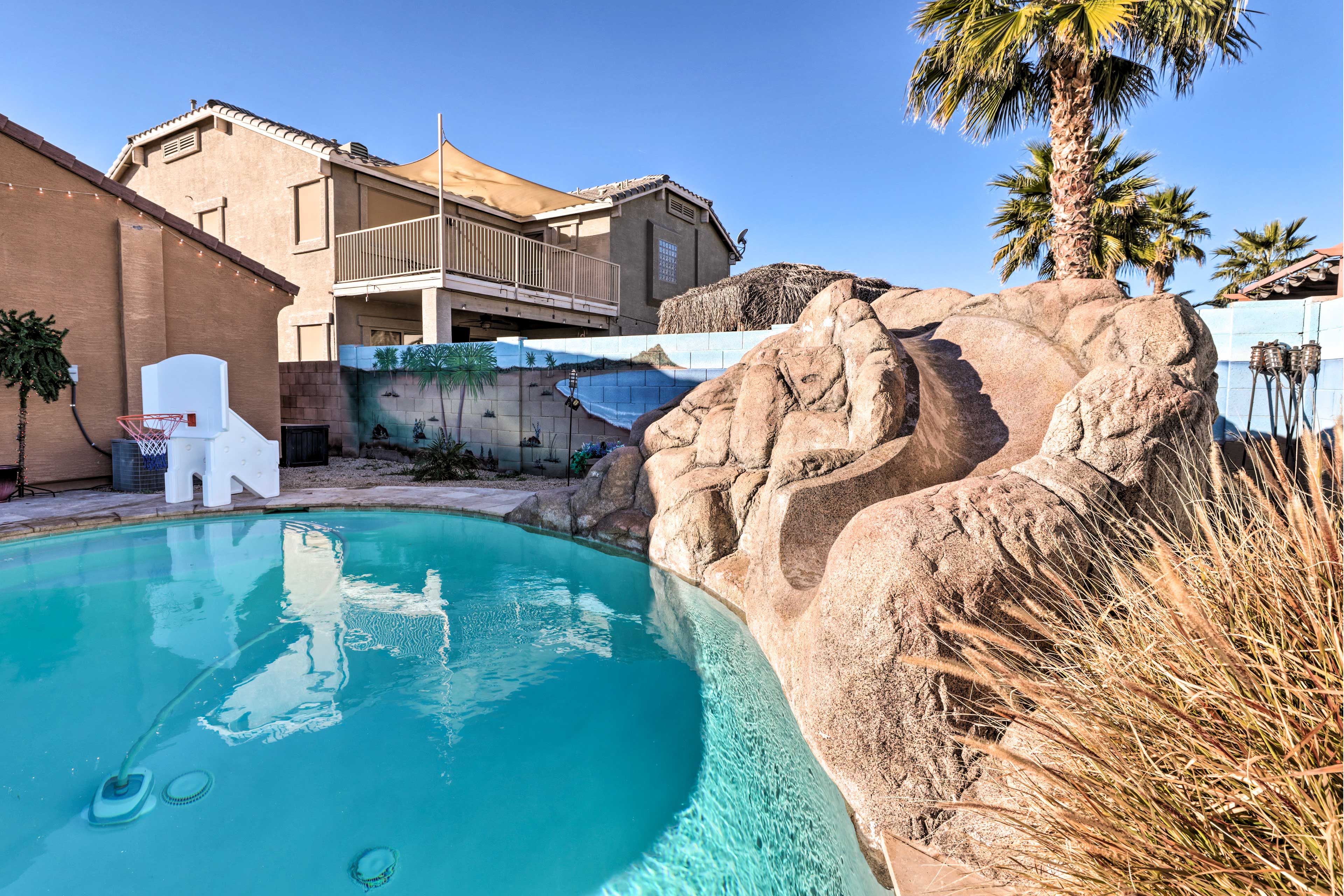 Une piscine avec une formation rocheuse, une chaise blanche et un bâtiment doté d’un balcon.