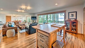 A spacious living room with a wooden dining table, leather sofas, and a large window offering a view of the outdoors. - Mid-century Cabin w/ Deck & Puget Sound Vistas! (Hansville)