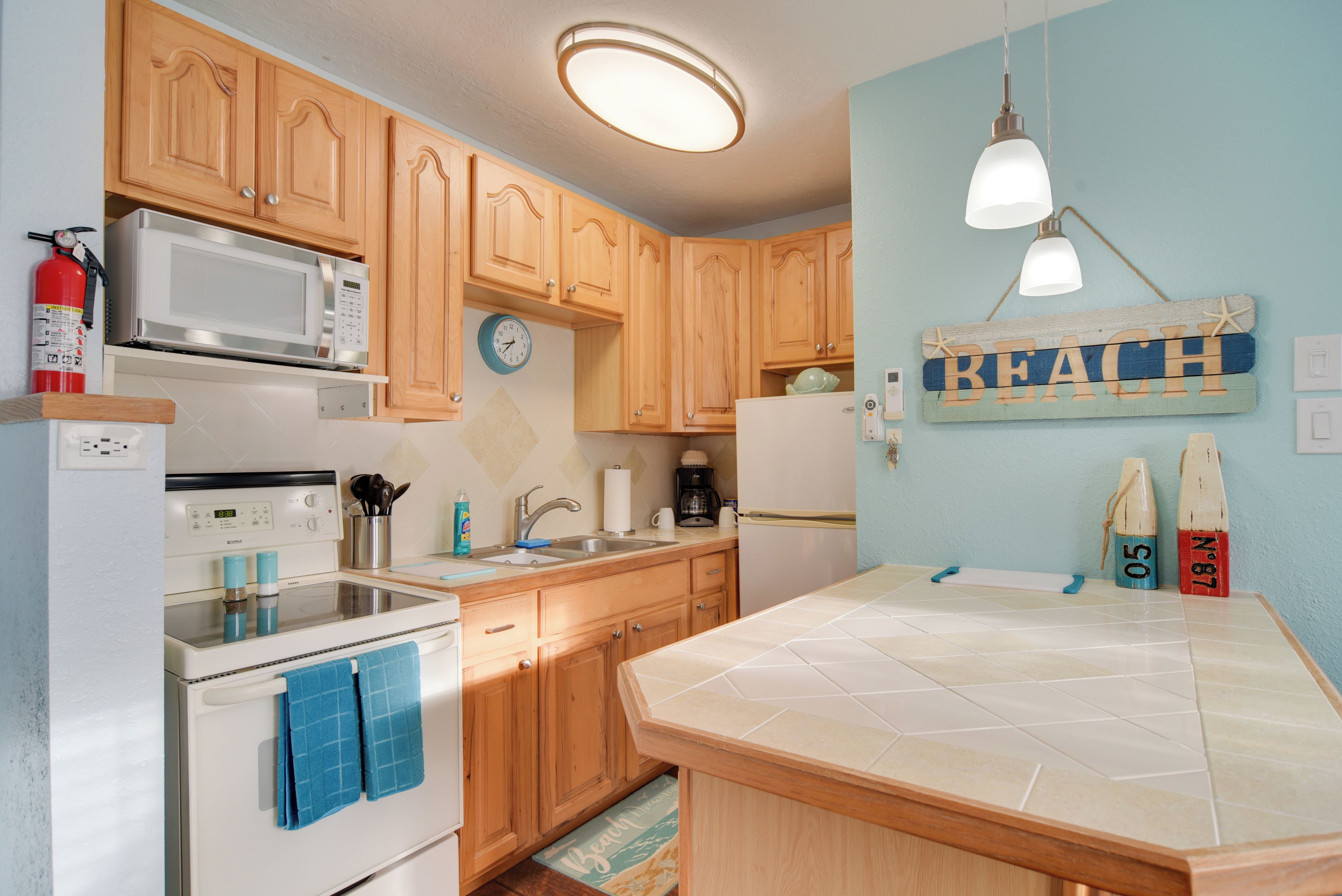 A kitchen with wooden cabinets, a microwave, a stove, and a sink. There is a clock on the wall and a sign that says "BEACH".