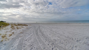 Une plage de sable où l’on voit des traces de pneus menant à l’eau, une jetée au loin et un ciel nuageux.