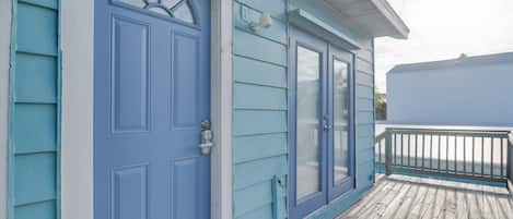 A blue house with a front door, a window, and a wooden deck.