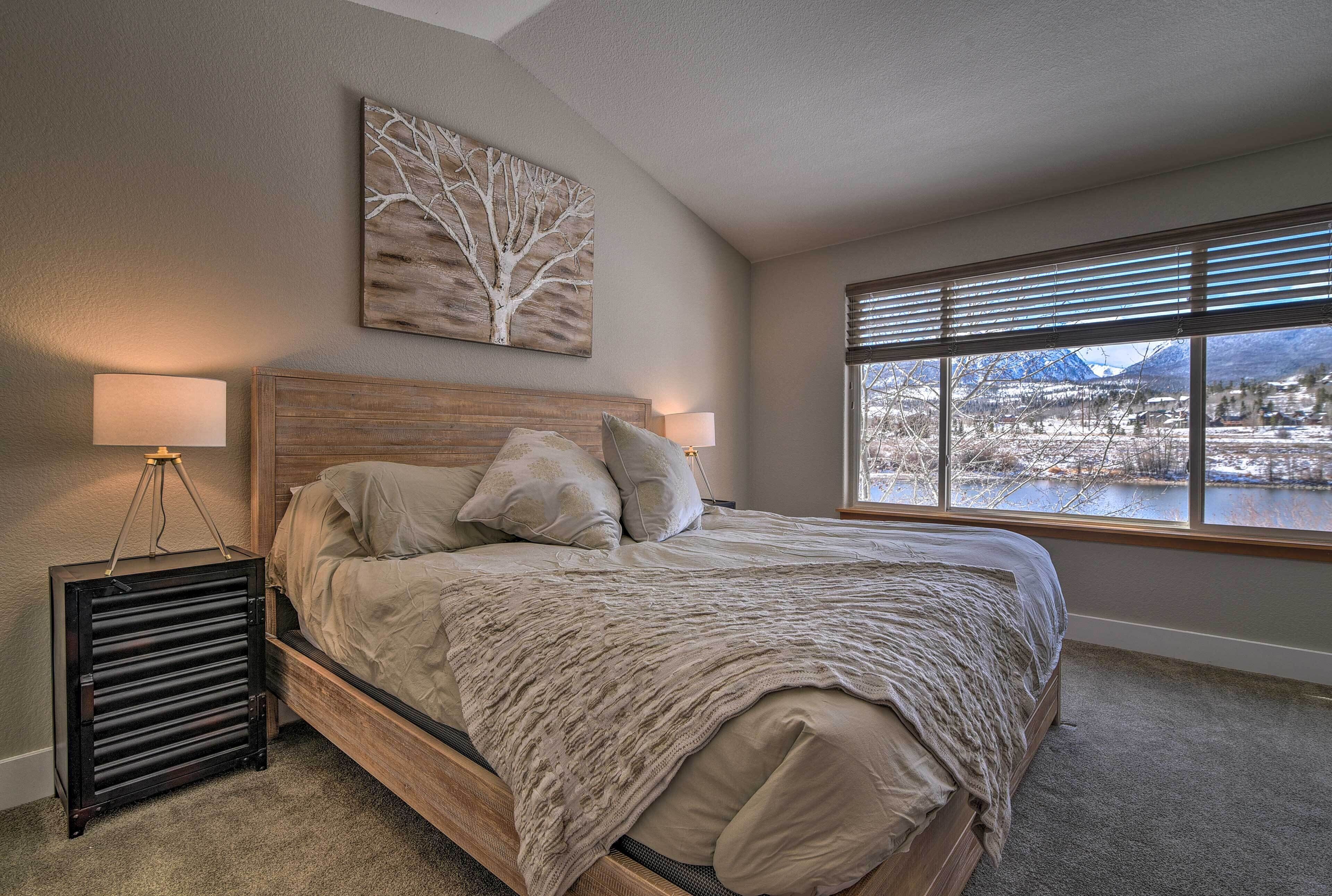 A bedroom with a bed, a nightstand, a lamp, a window with a view of a lake and mountains, and a wall art piece.