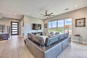 A spacious living room with a large grey sectional sofa, a flat-screen TV mounted on the wall, and a large window offering a view of the outdoors. - Fountain Hills Escape w/ Panoramic Mtn Views! (Fountain Hills)