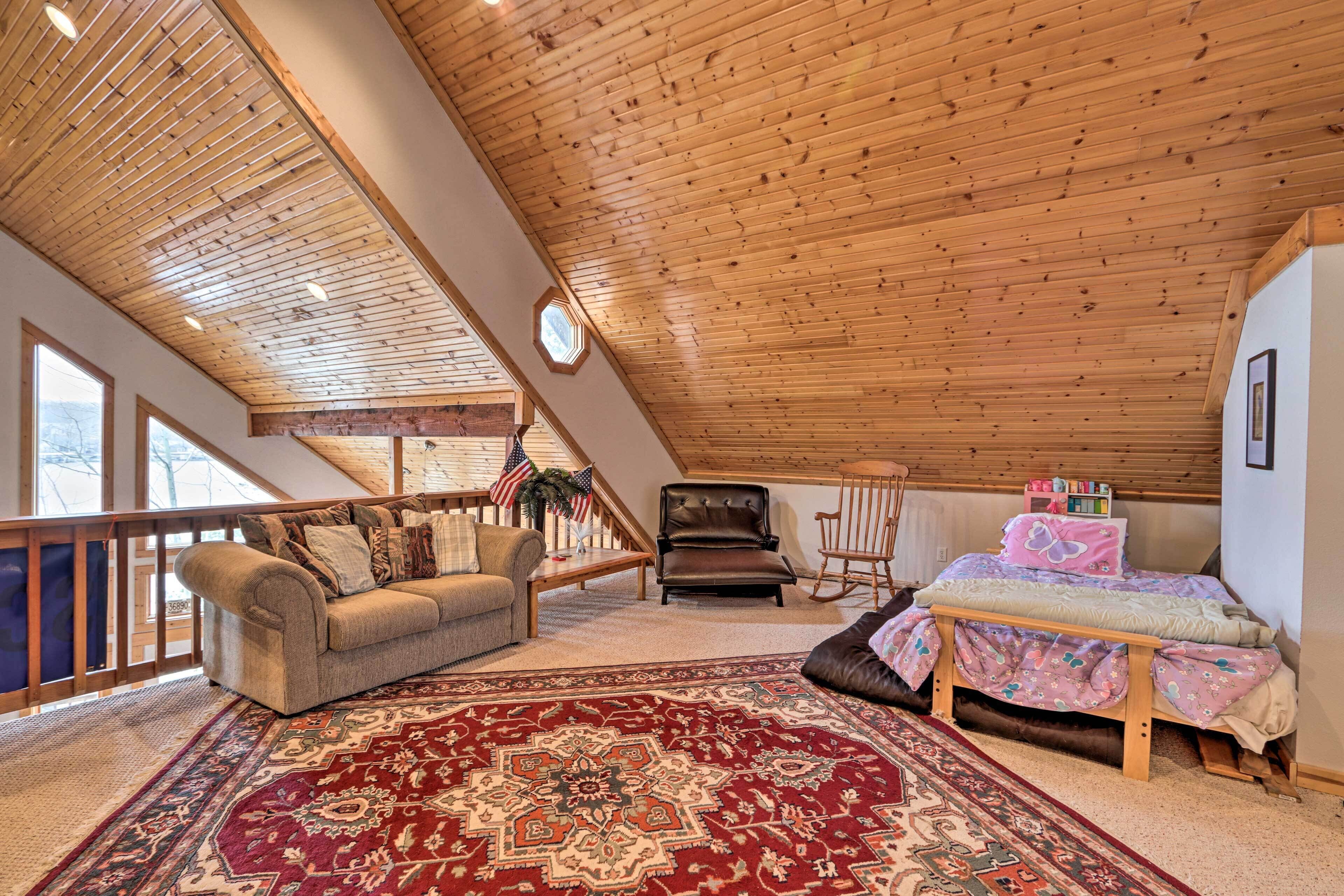 A cozy loft bedroom with a sloped ceiling, a large window, a wooden bed, a sofa, and a rocking chair.