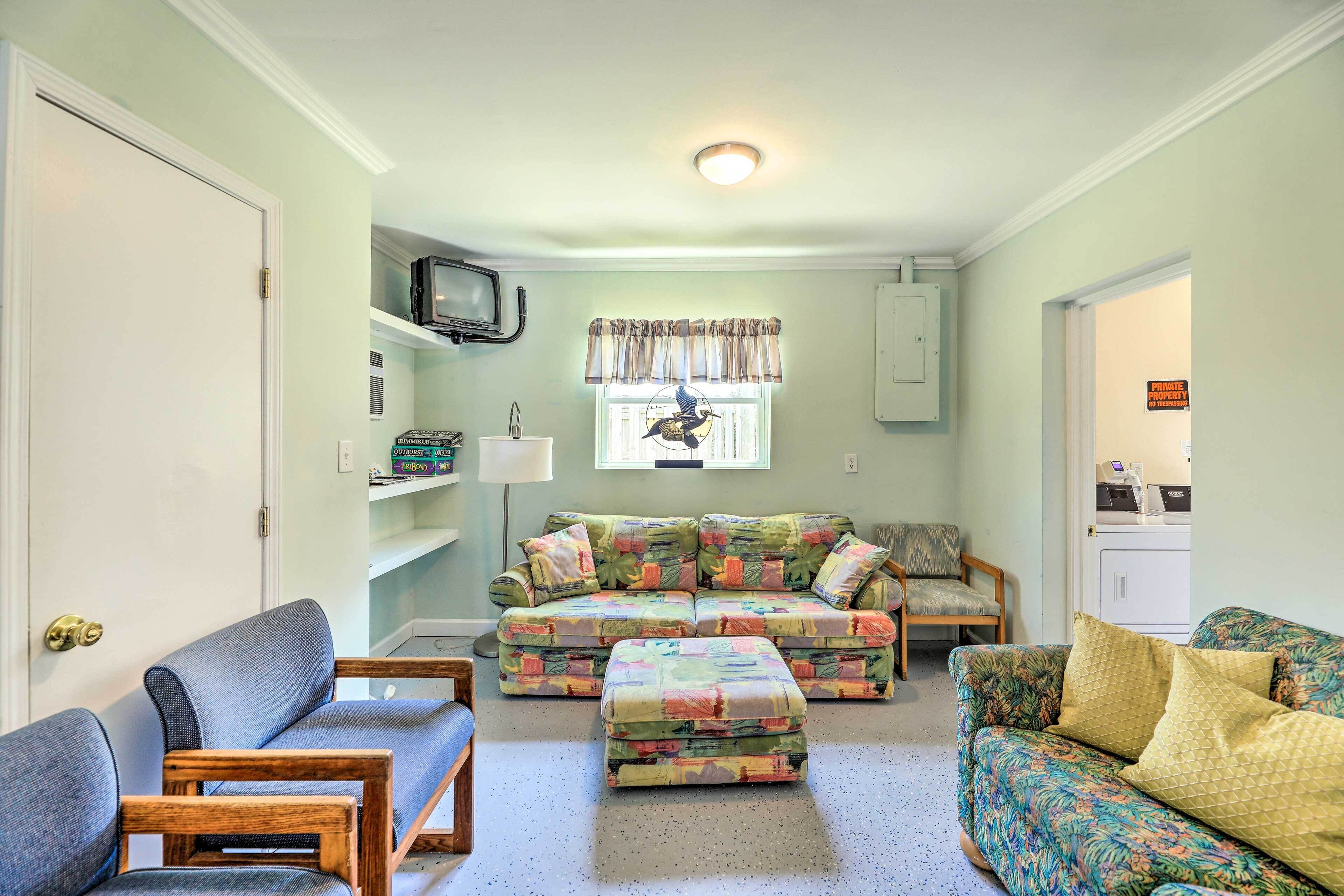 A living room with a sectional sofa, a loveseat, and a chair. There is a television on a shelf, a window with curtains, and a door with a sign.