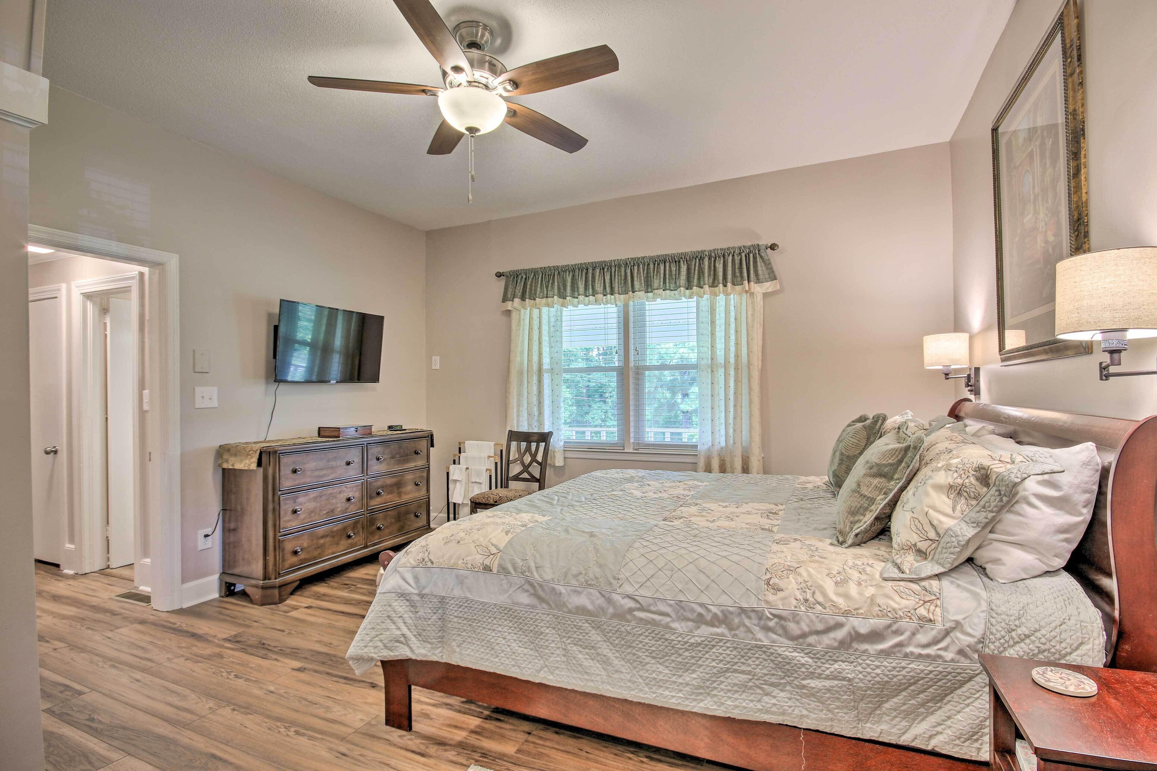 A bedroom with a wooden floor, a bed with a quilt, a ceiling fan, a television, a dresser, and a window with curtains.