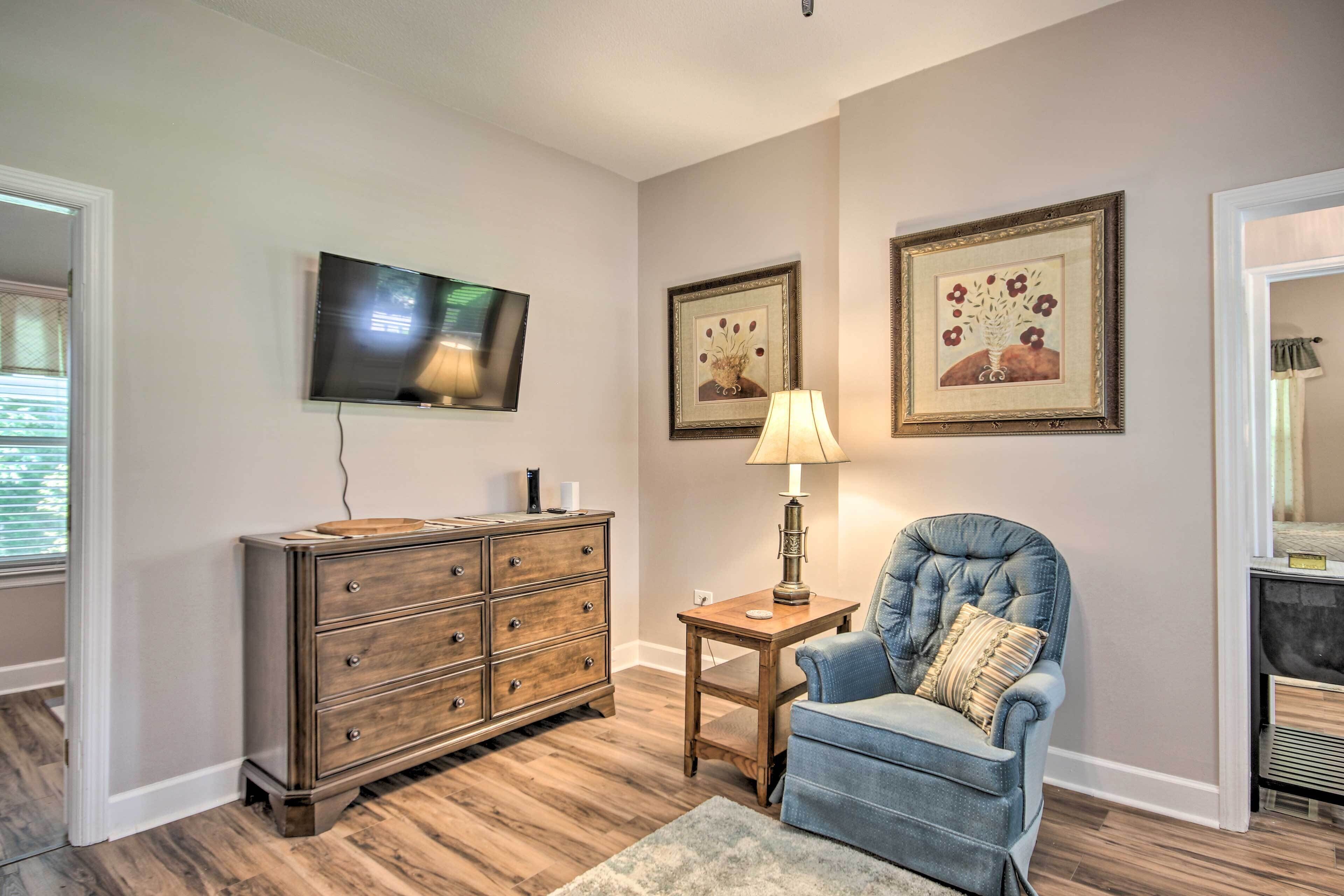 A room with a wooden dresser, a blue armchair, a small table with a lamp, and two framed pictures on the wall.