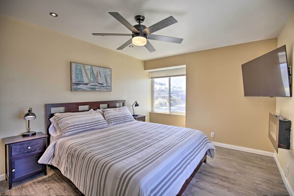 A bedroom with a bed, a nightstand, a ceiling fan, a painting, a window, and a flat-screen TV mounted on the wall. - Stunning Oceanside Condo: Walk to the Beach! (Oceanside)